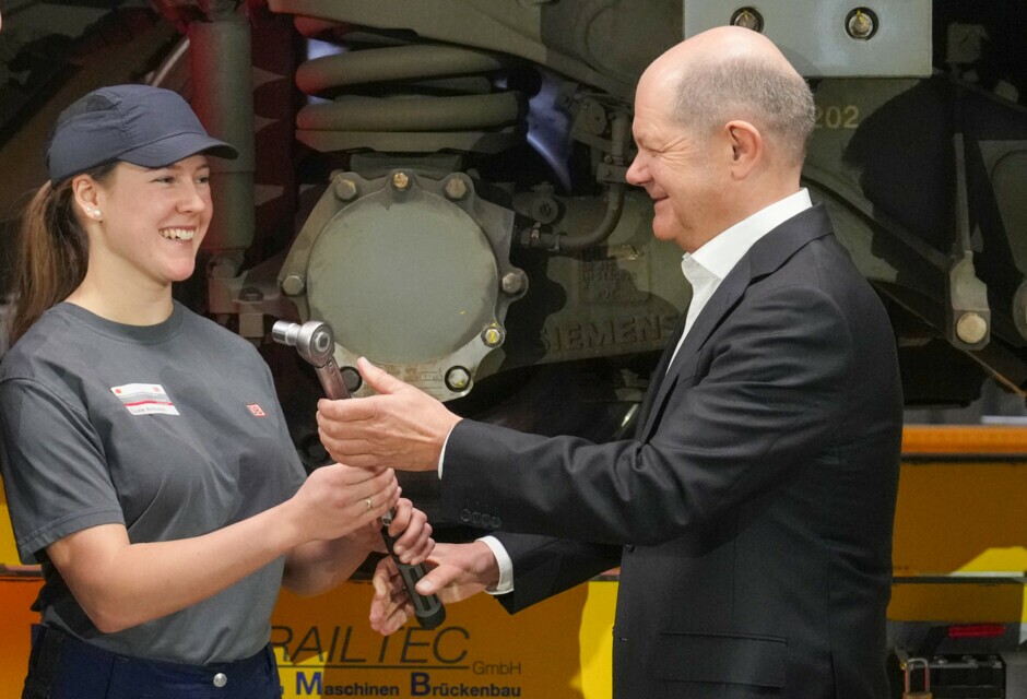 Bundeskanzler Olaf Scholz bekommt von einer Mitarbeiterin einen Schraubenschlüssel überreicht.
